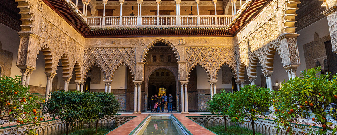 Tourists in the yard of Real Alcazar Royal Palace in Seville, Spain. The  Alcázar of Seville was founded as a fort in the 10th century by the Umayyad Caliphate on the site of a Roman fortress. Fly from London Luton Airport to Seville in 2 hours 40 minutes.